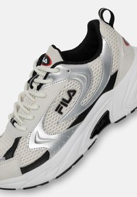 Scarpa atletica bianca e argento con pannelli in rete, dettagli neri, punta arrotondata e suola bianca spessa con grip testurizzato. Logo Fila presente.