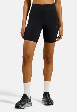 Femme portant un short de sport noir taille haute, des chaussettes blanches montantes et des chaussures de course noires, debout avec les bras détendus le long du corps.