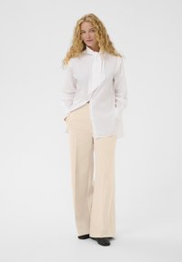 Chemise blanche à col cravate long, associée à un pantalon large beige ; le mannequin se tient avec les mains dans les poches, portant des chaussures noires.