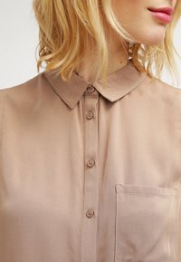 Blouse beige clair à col avec boutons sur le devant, une poche poitrine unique et une texture lisse. Le tissu semble léger et respirant.