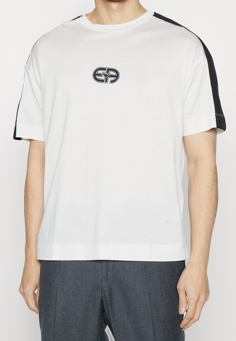 Vit bomulls t-shirt med korta ärmar och rund halsringning. Svarta ribbade detaljer på axlarna och en logotypapplikation på bröstet.