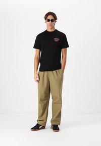 Black cotton t-shirt with red graphic, khaki baggy pants, black sneakers with gum soles, and dark sunglasses worn by a standing person.