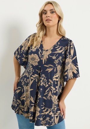 Lyshåret kvinde iført en løs marineblå bluse med beige blomster og lyseblå jeans, poserer med den ene hånd på hoften.