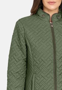 Chaqueta verde acolchada con un cuello alto, que presenta un cierre de cremallera y detalles de botones a presión. Patrón texturizado con bolsillos laterales y una silueta recta.