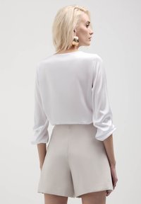 Blusa bianca semi-trasparente con maniche a tre quarti, abbinata a pantaloni corti beige a vita alta. Presenta una texture liscia e una vestibilità rilassata.