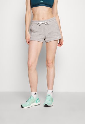 Kobieta w szarych szortach ze ściągaczem, turkusowym staniku sportowym, białych skarpetkach Adidas i miętowozielonych butach Adidas stoi na jednolitym białym tle.