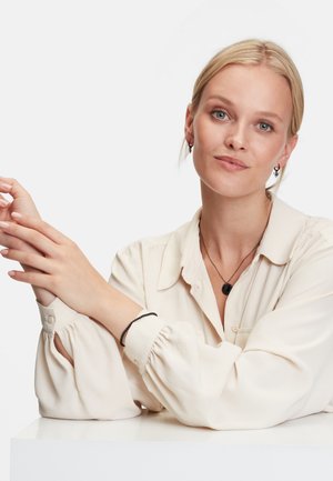 Beige Bluse mit Puffärmeln, mehrschichtige schwarze Halskette mit Herzanhänger, schwarzes Armband und Ohrringe mit Hoop-Design. Hände auf einer weißen Fläche gefaltet.