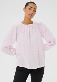 Blouse rose clair, transparente, avec des pois texturés, des manches longues bouffantes et un col rond. Pantalons noirs ajustés portés en dessous.