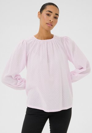 Blouse rose clair, transparente, avec des pois texturés, des manches longues bouffantes et un col rond. Pantalons noirs ajustés portés en dessous.