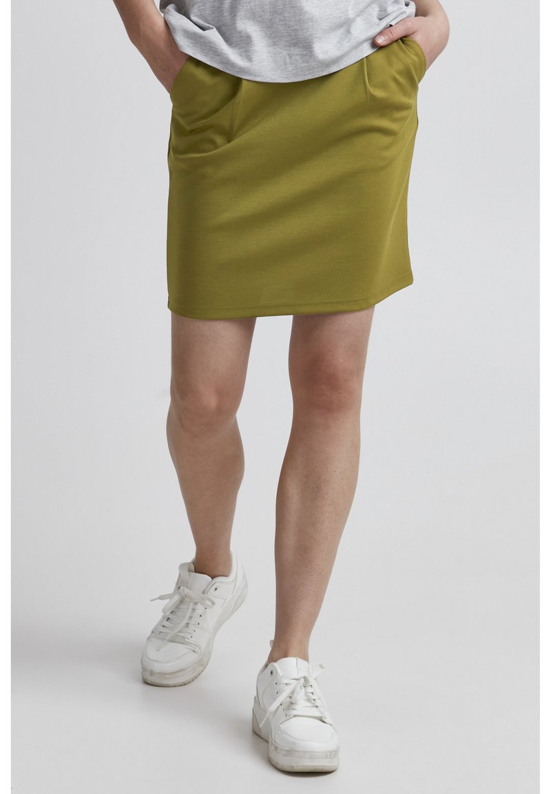 Jupe olive en tissu doux, dotée de deux poches latérales, d'une coupe droite et d'une finition au-dessus du genou, assortie de baskets blanches.