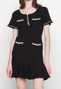 Schwarzes geripptes Kleid mit kurzen, rüschendetails Ärmeln. Verfügt über einen gewellten Ausschnitt, zwei vordere Taschen mit kontrastierendem Besatz und einen ausgestellten Saum.
