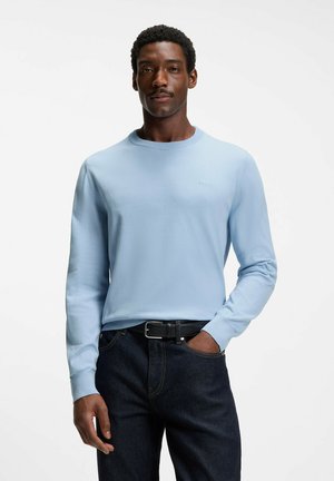 Man standing with one hand in pocket, wearing light blue crewneck sweater and dark jeans against plain white background.