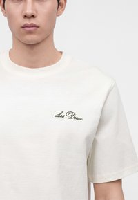 Νέος άνδρας που φοράει ένα απλό λευκό T-shirt με μικρό, σκούρο πράσινο κείμενο "Les Deux" τυπωμένο στο αριστερό στήθος σε γκρι φόντο.