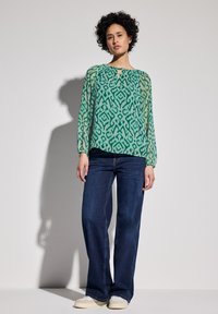 Grüne gemusterte Bluse mit langen Ärmeln, weichem Stoff und rundem Ausschnitt, kombiniert mit dunkelblauen weiten Jeans und hellen Schuhen.