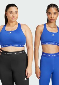 Two women in fitted blue sports bras and black or blue leggings, showcasing a smooth, stretchy fabric with contrasting branded waistbands.