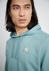 Felpa leggera color turchese con cappuccio e coulisse, e un patch bianco con il logo Puma sul petto. Tessuto morbido e liscio con una vestibilità comoda.