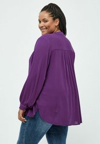 Blusa viola con maniche lunghe e leggermente a sbuffo, polsini con bottone e pieghe sul retro. L'orlo ha una forma curvata ed è abbinata a jeans blu.