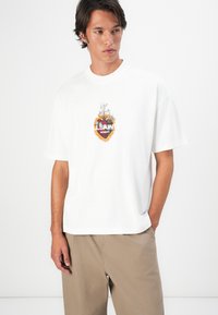 Jonge man in een wit T-shirt met een vlammenhartgrafiek en de tekst "LSAINTS LONDON", gecombineerd met beige broek, staand tegen een witte muur.