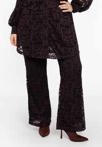 Zwarte kanten wijde-pantalon gecombineerd met een bijpassende lange kanten tuniek en bruine puntige hoge hakken op een staand persoon.