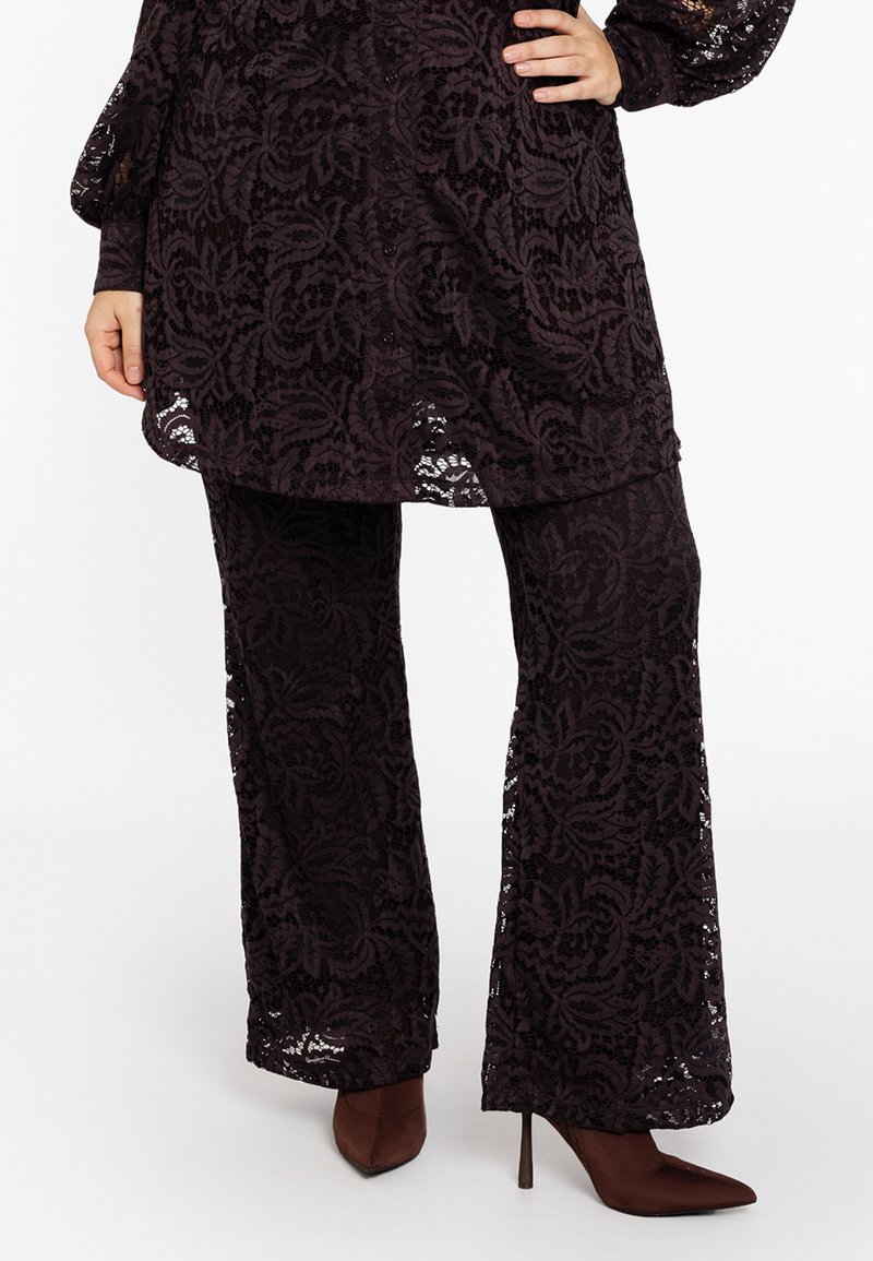 Zwarte kanten wijde-pantalon gecombineerd met een bijpassende lange kanten tuniek en bruine puntige hoge hakken op een staand persoon.