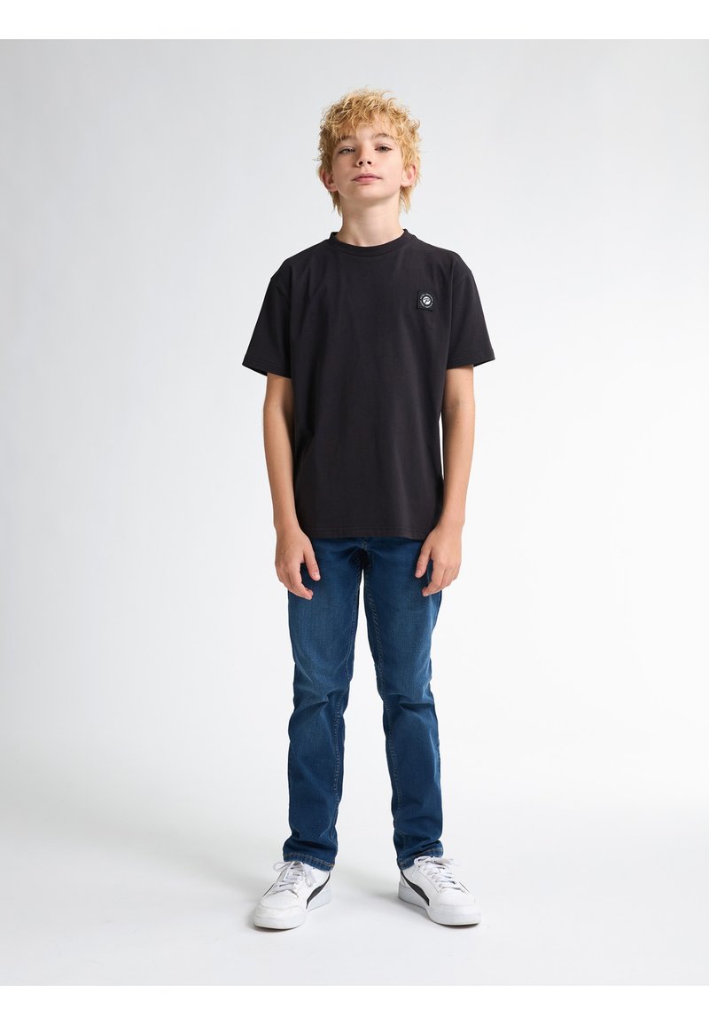 Černé bavlněné tričko s kulatým výstřihem a malou nášivkou loga na hrudi, spárované s modrými džíny a bílými teniskami.