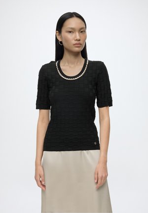 Femme aux cheveux noirs et raides, portant un haut noir texturé à manches courtes avec un détail perlé au niveau du col et une jupe beige, debout sur fond blanc.