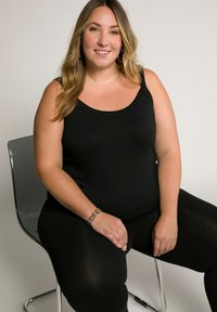 Černý bezramínkový top a černé legíny. Modelka má na sobě barevný korálkový náramek. Sedí na průhledné židli na světlém pozadí.