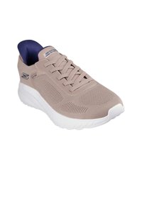 Beżowy but sportowy z tkaninową cholewką, sznurowanym zapięciem i białą gumową podeszwą. Charakteryzuje się teksturowaną powierzchnią oraz wyściółką w okolicy kostki.