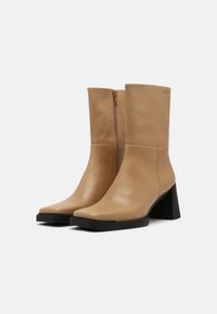 Beżowe skórzane botki za kostkę z kwadratowym noskiem i słupkowym obcasem. Posiadają boczny zamek błyskawiczny i gładką fakturę. Czarna gumowa podeszwa.