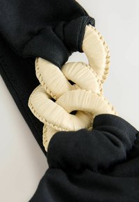Gros plan sur des anneaux tissés beiges entrelacés, reliés par des boucles de tissu noir sur une surface textile noire texturée.