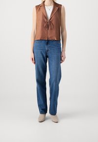 Brun linnen ärmlös topp med krage och knytdetalj, i kombination med blå raka jeans och ljusbeige ankelstövlar.