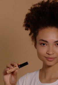 Tube de correcteur KIKO beige avec bouchon noir tenu dans la main. Texture lisse, design minimaliste. Le modèle porte un haut blanc orné de perles.