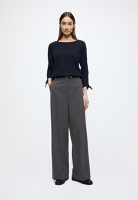 Granatowa teksturowana bluzka z długim rękawem w połączeniu z szarymi spodniami o szerokich nogawkach. Czarny pasek z klamrą i ciemne buty dopełniają strój.