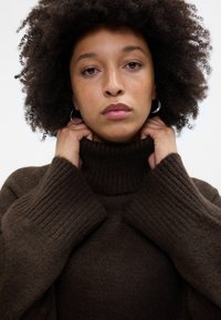 Brūns ribots augstais kakls džemperis ar mīkstu tekstūru. Modelis tur apkakli, demonstrējot pārāk lielas piedurknes un piegulošu dizainu.