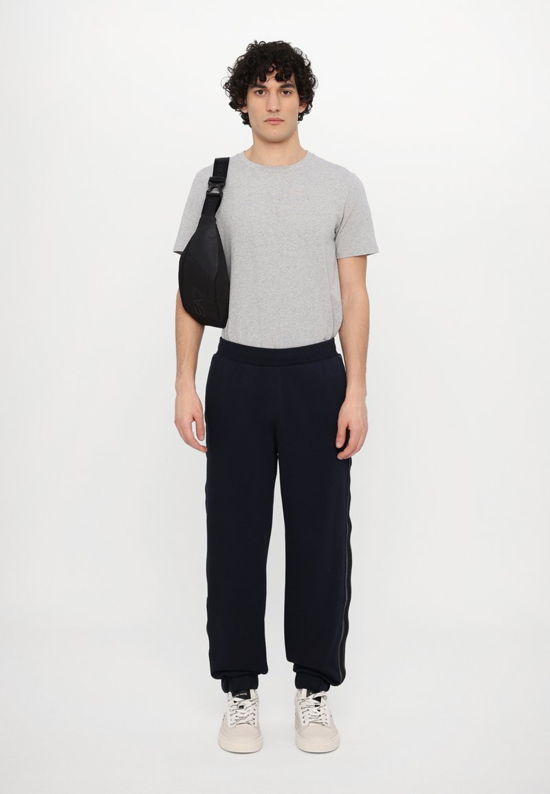 T-shirt grigio a maniche corte, pantaloni della tuta blu navy con strisce laterali e sneakers bianche. Una borsa a tracolla nera è indossata sulla spalla sinistra.