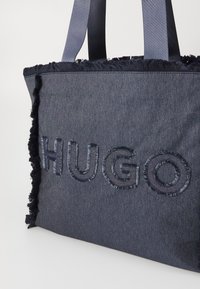 Tamsiai mėlyna džinsinė rankinė su pluoštiniais kraštais, ant kurios šiek tiek šviesesniu mėlynu siuvinėta "HUGO". Tvirtas audinys ir dvi rankenos.