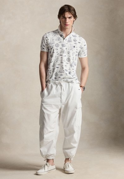 Νέος άνδρας με λευκό πόλο με ναυτικά σχέδια, λευκό cargo παντελόνι με κορδόνια στους αστραγάλους και λευκά αθλητικά παπούτσια, στέκεται με τα χέρια στις τσέπες.