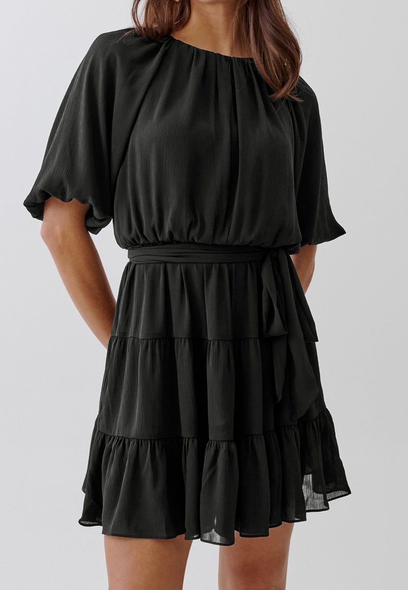 Femme portant une courte robe noire avec des manches bouffantes, une taille froncée, une jupe à étages et une ceinture en tissu nouée sur le côté.