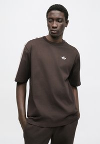 Κοντομάνικο καφέ t-shirt από βαμβάκι, με στρογγυλή λαιμόκοψη, διακριτικό λογότυπο στο στήθος και λευκές ρίγες στους ώμους.