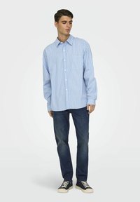 Hellblau-weiß gestreifte Button-down-Bluse mit langen Ärmeln und einer Brusttasche, kombiniert mit dunklen Jeans und schwarzen Sneakern.