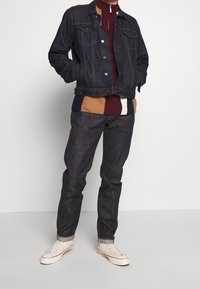 Chaqueta de mezclilla oscura con bolsillos en el pecho, usada sobre un suéter a bloques de color en tonos marrones y burdeos. Combinada con jeans oscuros y zapatillas blancas.