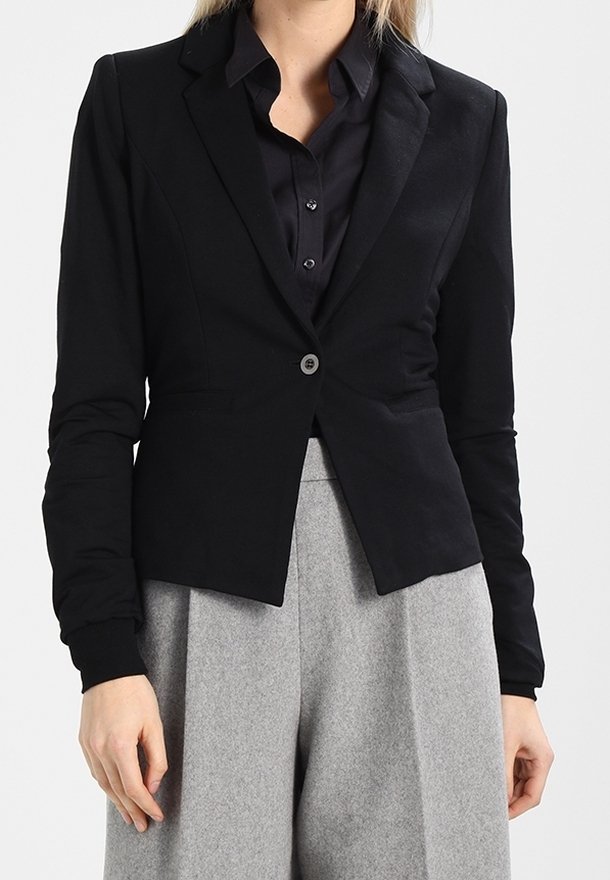 Femme portant un blazer noir ajusté sur une chemise noire boutonnée et un pantalon gris taille haute à jambes larges sur un fond uni.