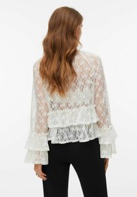 Blusa blanca de encaje con volantes en capas, mangas largas y diseño transparente. La parte trasera muestra una tela transparente con detalles de encaje intrincados.