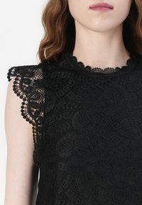 Svart blonde topp med intrikate blomster mønstre, bølgete skulderdetaljer og en teksturert overflate. Rund utringning med detaljerte kanter.