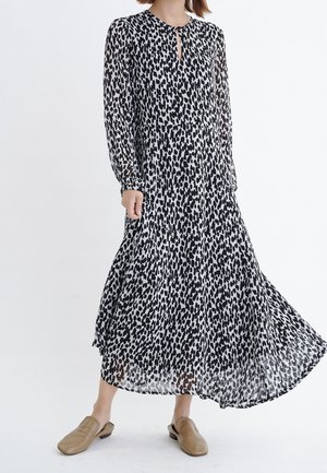 Femme portant une longue robe à imprimé léopard noir et blanc avec des manches longues et des chaussures beige sans lacets, debout sur un fond blanc uni.