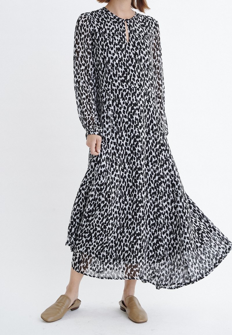 Femme portant une longue robe à imprimé léopard noir et blanc avec des manches longues et des chaussures beige sans lacets, debout sur un fond blanc uni.