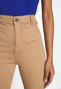 Ljusbruna högmidjade jeans med en slät textur, som har en silverfärgad knappstängning. Tyget är stretchy och sitter åtsittande runt låren.