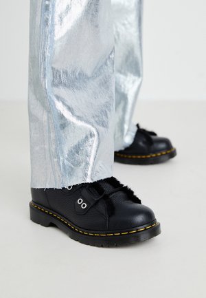 Chaussures en cuir noir avec couture jaune, doublure en fourrure et œillets argentés portées avec un pantalon argenté brillant sur un sol blanc.
