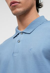 Világoskék pamut póló, gallérral, két gombos felsőrésszel és egy kis hímzett logóval a mellkason.