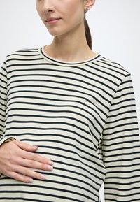 Langermet skjorte med kremfarget bakgrunn og tynne marineblå striper, rund halslinning og myk stofftekstur. Hånd plassert på magen.